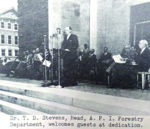 Man stands at podium in front of seated colleagues on the steps of the Forestry Building, circa 1949.