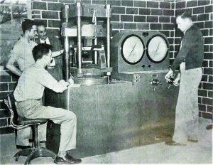Student tests wood in 100,000 pounds tester in timber physics laboratory, circa 1949. One student operates the machine while two others record with an instructor observing.