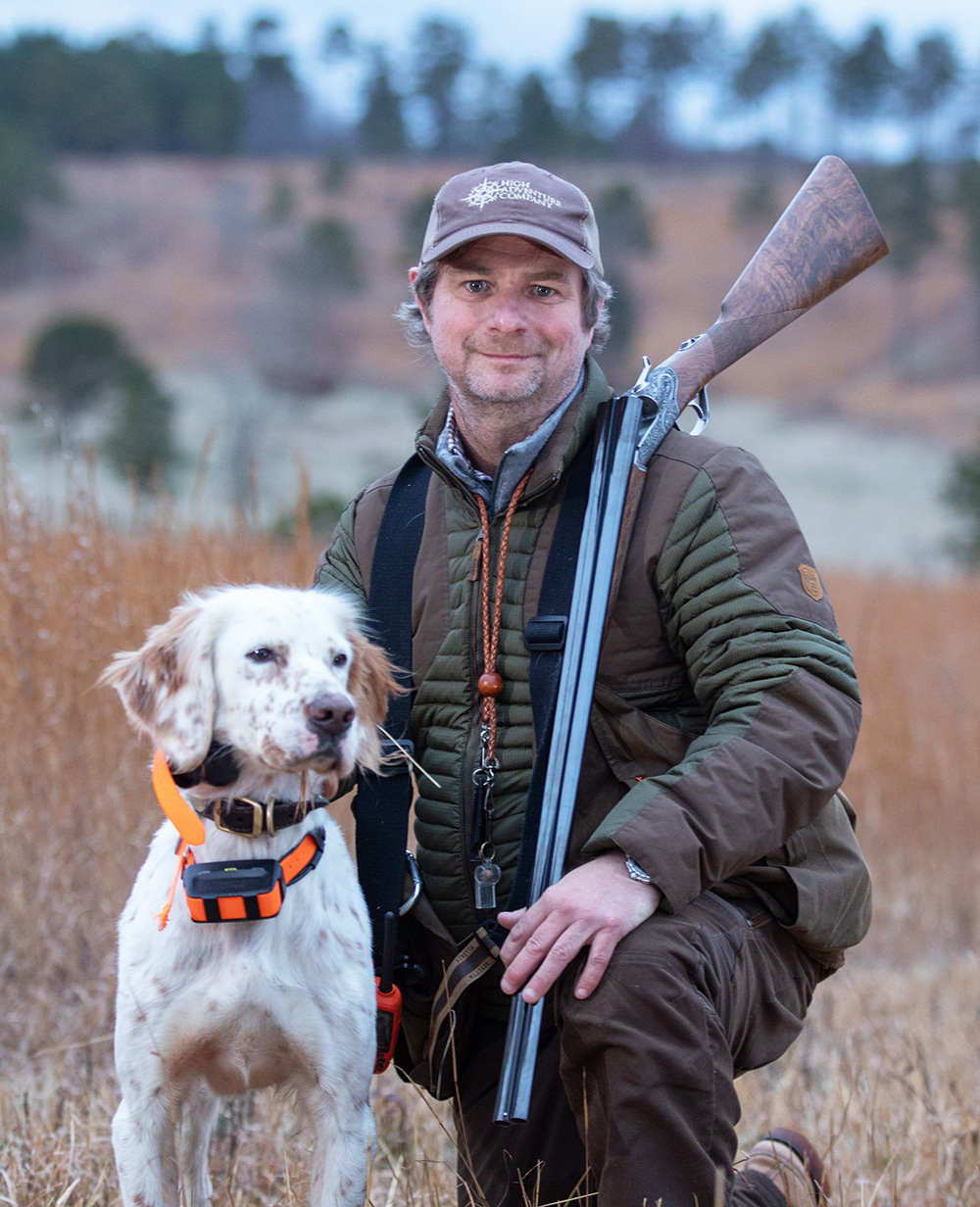 John Burrell kneeling outdoors in grassy field with hunting dog.
