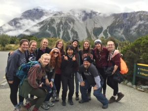 Dr. Morse crouches with a group of students in front of a cloudy mountain range and forest in New Zealand.