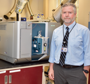 Dr. Jason White stands in a lab in front of a blue and silver machine. He wears a light blue button down, a striped black tie, a name tag on a lanyard and gray slacks.