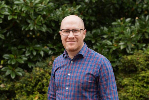 Man in blue shirt standing in front of green tree.