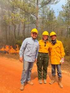 Three people wearing yellow hard hats and fieldwork clothing stand smiling on a dirt road in a pine forest during a prescribed burn, with low flames and smoke visible behind them among the trees.