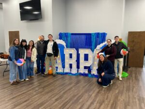 A group of people posing indoors beside large illuminated “ARPA” letters with a blue backdrop and beach-themed props.
