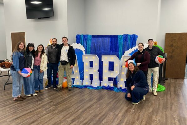 A group of people posing indoors beside large illuminated “ARPA” letters with a blue backdrop and beach-themed props.