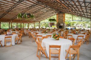 Crooked Oaks Pavilion set for an Auburn Athletics event with round tables, white linens, wooden chairs, and simple floral centerpieces, under string lights in an open-air space.