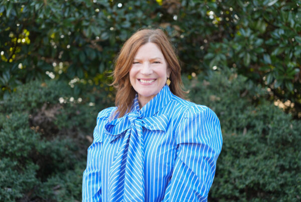 Woman with red hair in a blue shirt smile in front of a green tree