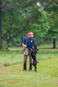 A man embraces his bird dog in a field with a shotgun broken over his shoulder.