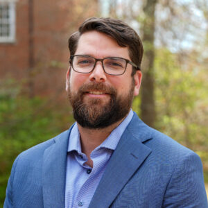 Man with glasses and a beard wearing a blue blazer, standing outdoors in front of trees and a brick building.
