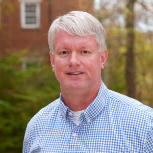 Man with gray hair wearing a blue checkered shirt, standing outdoors with trees and a brick building in the background.