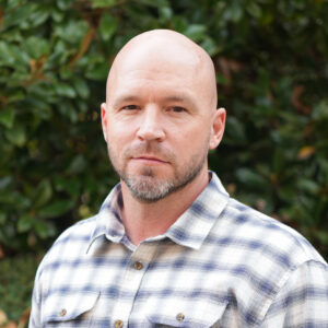 John Richards poses for a photo in a white plaid shirt against a green background.