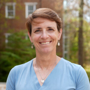 Woman with short brown hair wearing a light blue top and earrings, smiling outdoors in front of trees and a brick building.