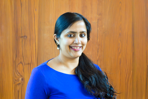 Woman with dark hair and in a blue shirt smiling against a medium toned wooden background.