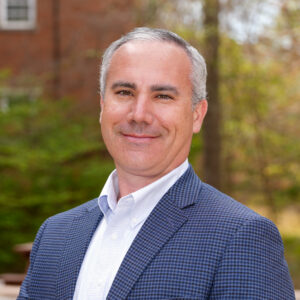 Man with short gray hair wearing a patterned blazer and white shirt, standing outdoors with trees and a brick building in the background.