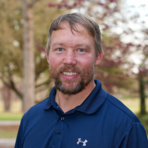 Man with short hair and a beard wearing a navy polo shirt, smiling outdoors with trees in the background.