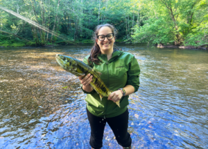 Person standing in a shallow river holding a fish, with trees and sunlight in the background.