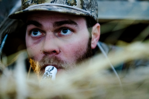 A hunter holds his lips to a duck call while peering up into the sky.