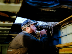 A hunting guide holds a waterfowl call to his mouth while looking upward out of a hunting blind.
