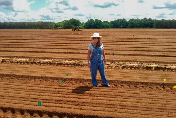 Nina Payne stands in a tree nursery with a tractor operating behind her.