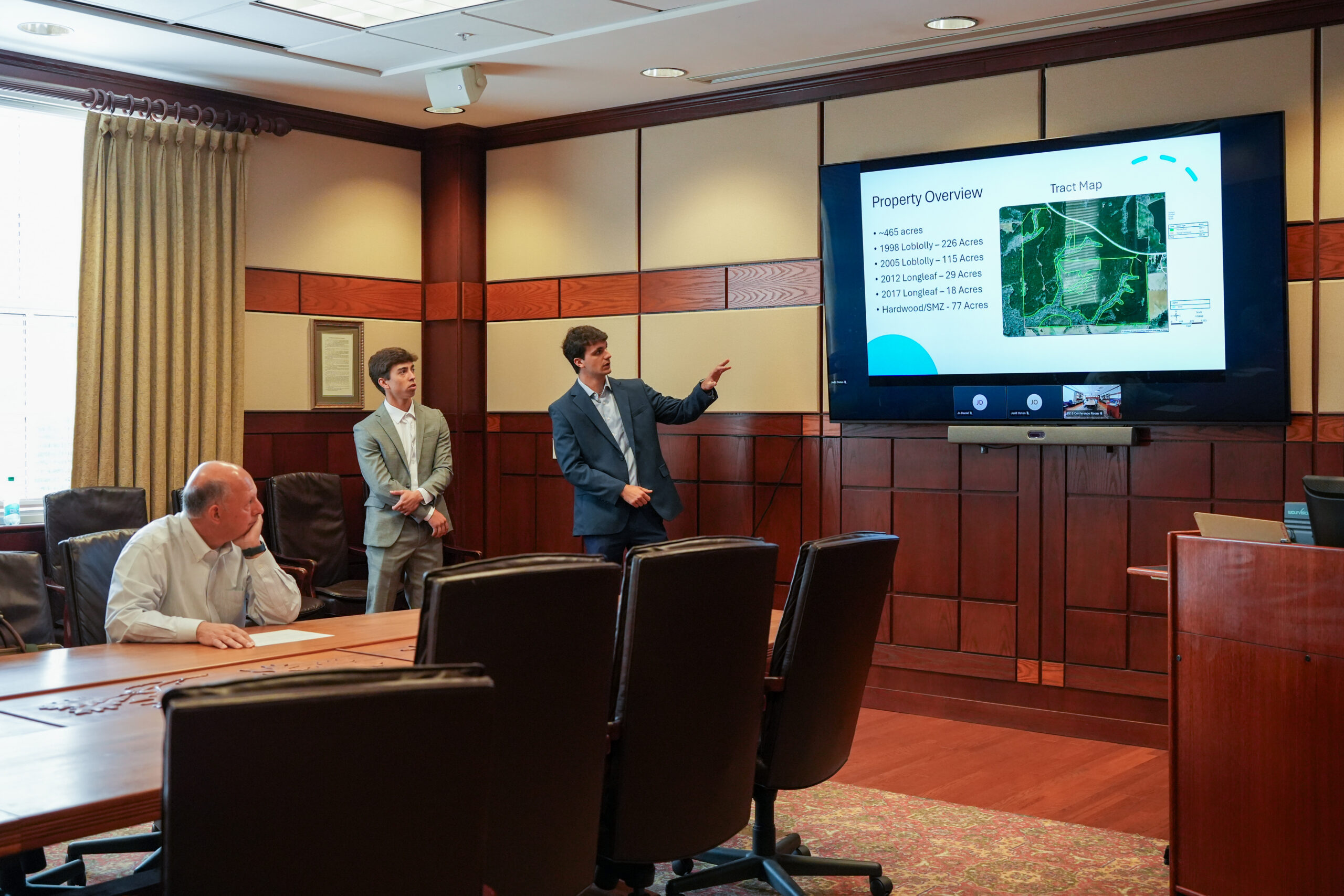 two male students give a presentation in a boardroom with one man seated
