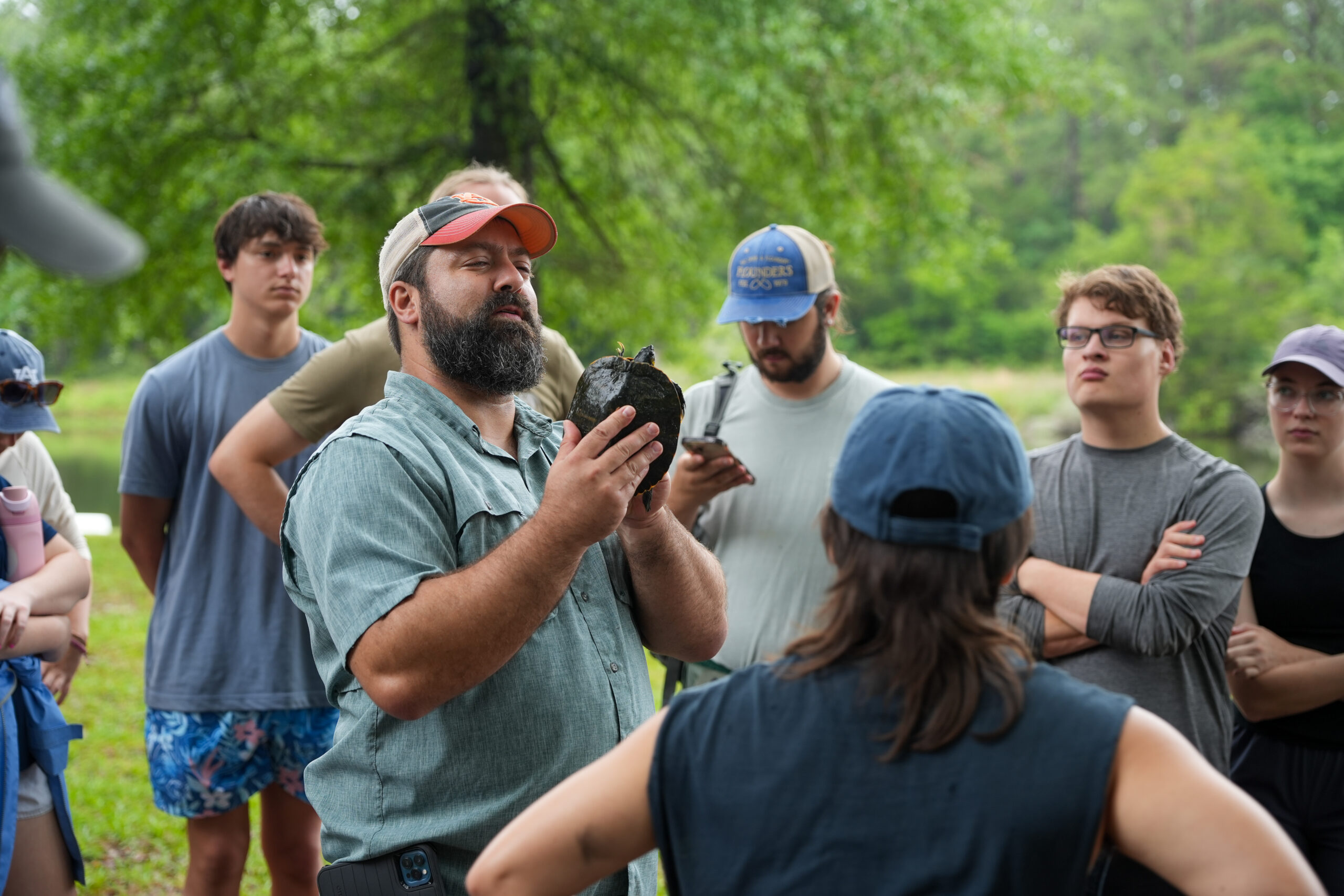 professor demonstrates handling and measurement of a turtle next to a pond with a group of students looking on