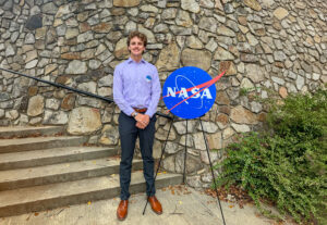 Sid Warrent stands next to a NASA emblem on an easel in front of a stone backdrop.