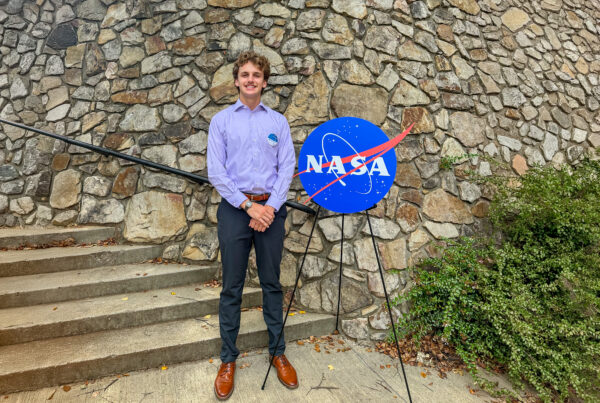 Sid Warrent stands next to a NASA emblem on an easel in front of a stone backdrop.