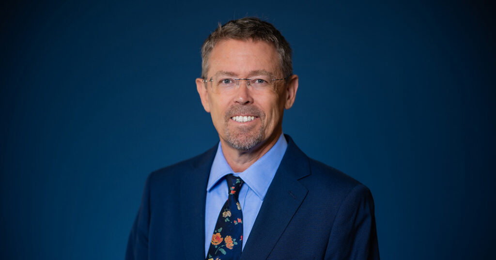 A person with short hair and glasses smiles while wearing a dark blue suit, light blue shirt, and a floral-patterned tie against a solid blue background.