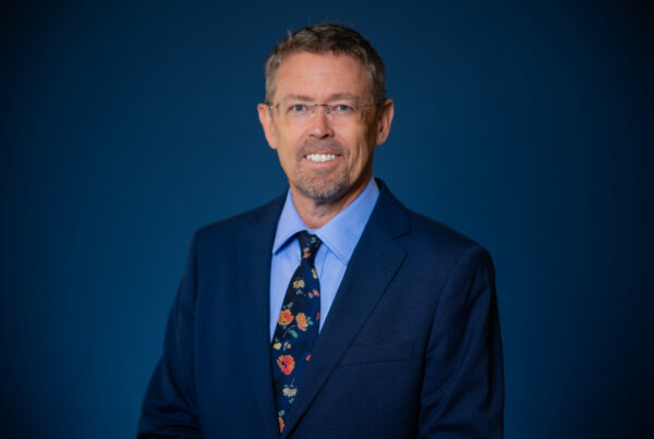 A person with short hair and glasses smiles while wearing a dark blue suit, light blue shirt, and a floral-patterned tie against a solid blue background.